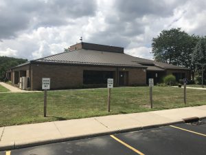 Small building at top of hill with large windows and clean sheet metal roof taken from parking lot with Visitor parking signs